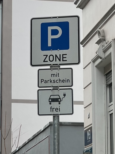 Ein Verkehrsschild aus mehreren Schildern. Oberes Schild „P-Zone“, mittleres Schild „mit Parkschein“ unteres Schild zeigt das Piktogramm eines Elektroautos mit der Unterschrift „frei“