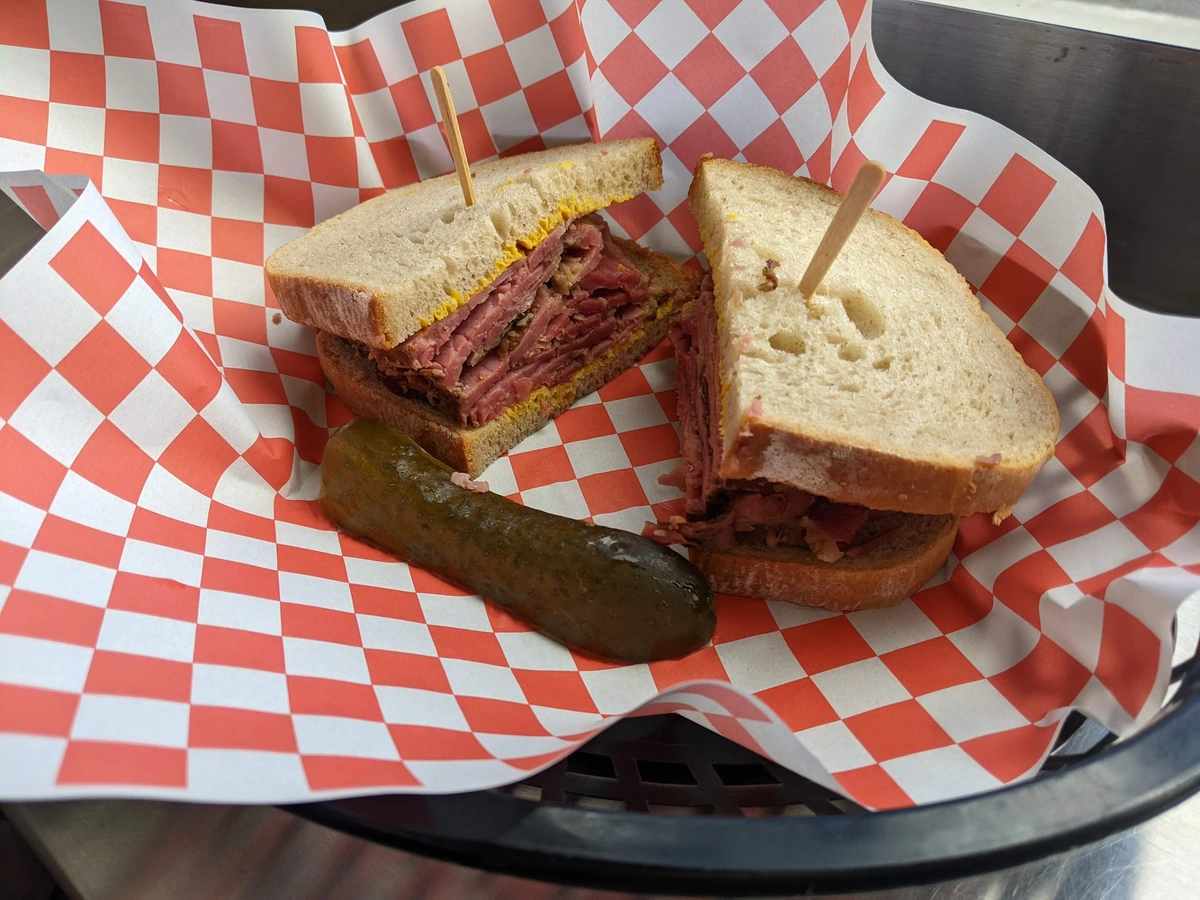A smoked meat sandwich, cut into two and with a toothpick in each half. It sits in a basket alongside a slice of pickle.